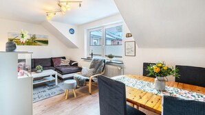 Ett modernt vardagsrum med en soffa, fåtölj, matbord och utsikt över omgivningen genom ett fönster.