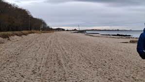 En sandstrand med spår av däck, en träbrygga och en avlägsen vy över en fyr.