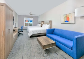 A hotel room with a bed, a blue sofa, a wooden coffee table, a desk with a chair, and a view of the outdoors through a window.