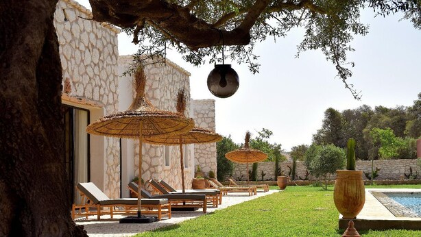 Zona de piscina con sillones de descanso y sombrillas, un edificio de piedra y un árbol.