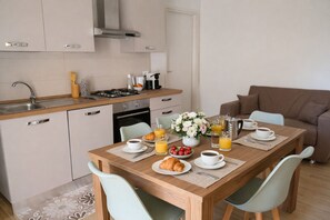 A modern kitchen with a dining area, featuring a wooden table set for breakfast with croissants, fruit, and beverages.
