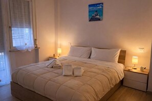 A neatly made bed with a beige quilt, two bedside tables with lamps, and a painting of a coastal scene on the wall.