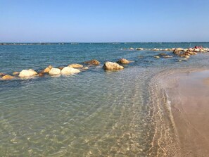 A beach with clear shallow waters, large rocks, and a sandy shore.
