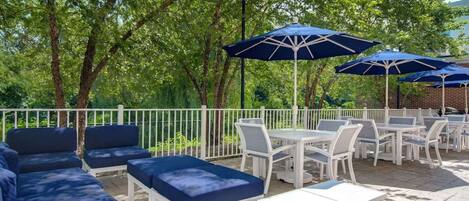 Une terrasse extérieure avec des meubles blancs et des coussins bleus, entourée d’arbres et ombragée par des parasols bleus.
