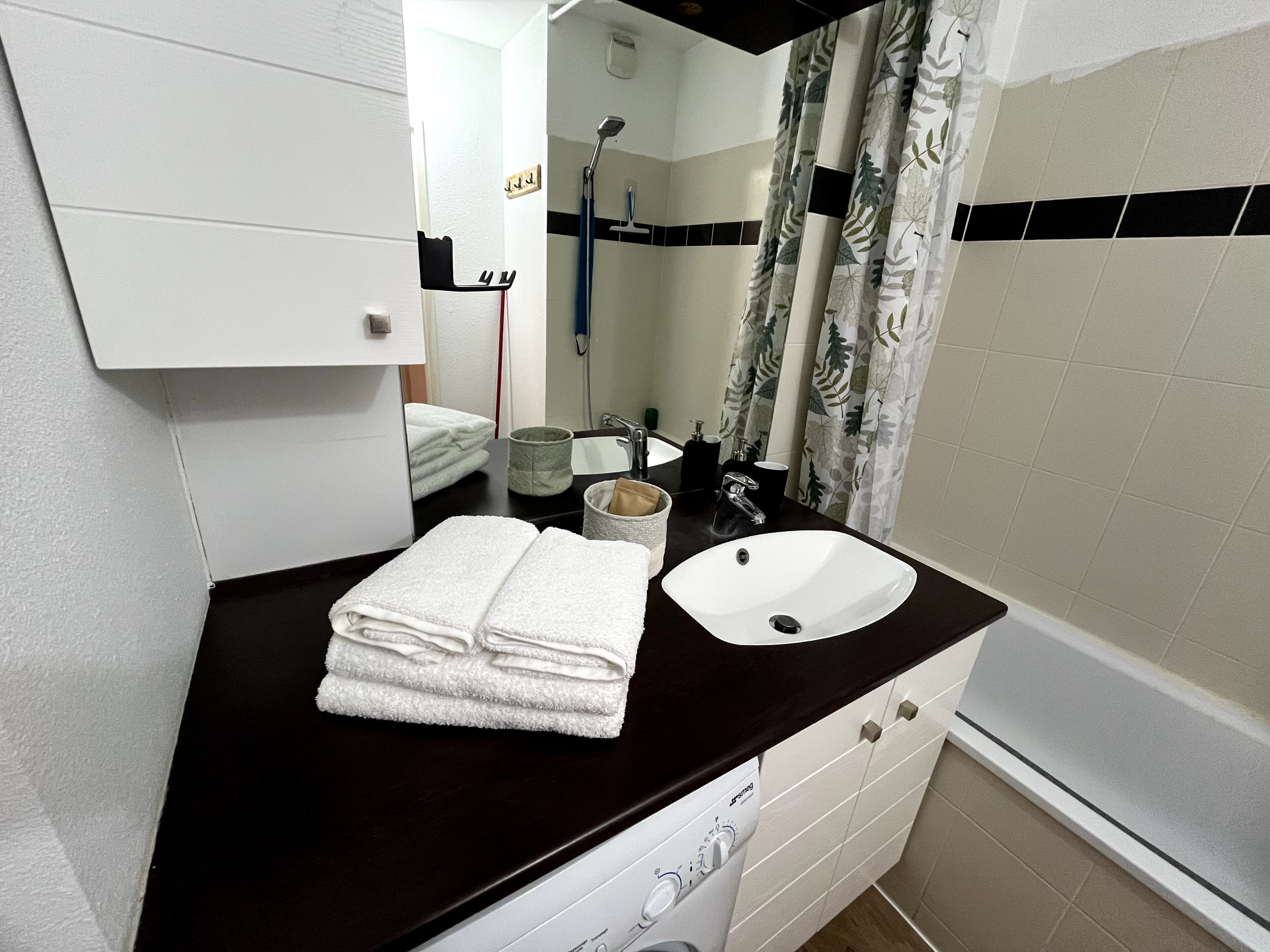 Een badkamer met een witte wastafel, een donker aanrecht en een spiegel erboven. Handdoeken liggen gestapeld op het aanrecht, en een douchekop is in de achtergrond zichtbaar.