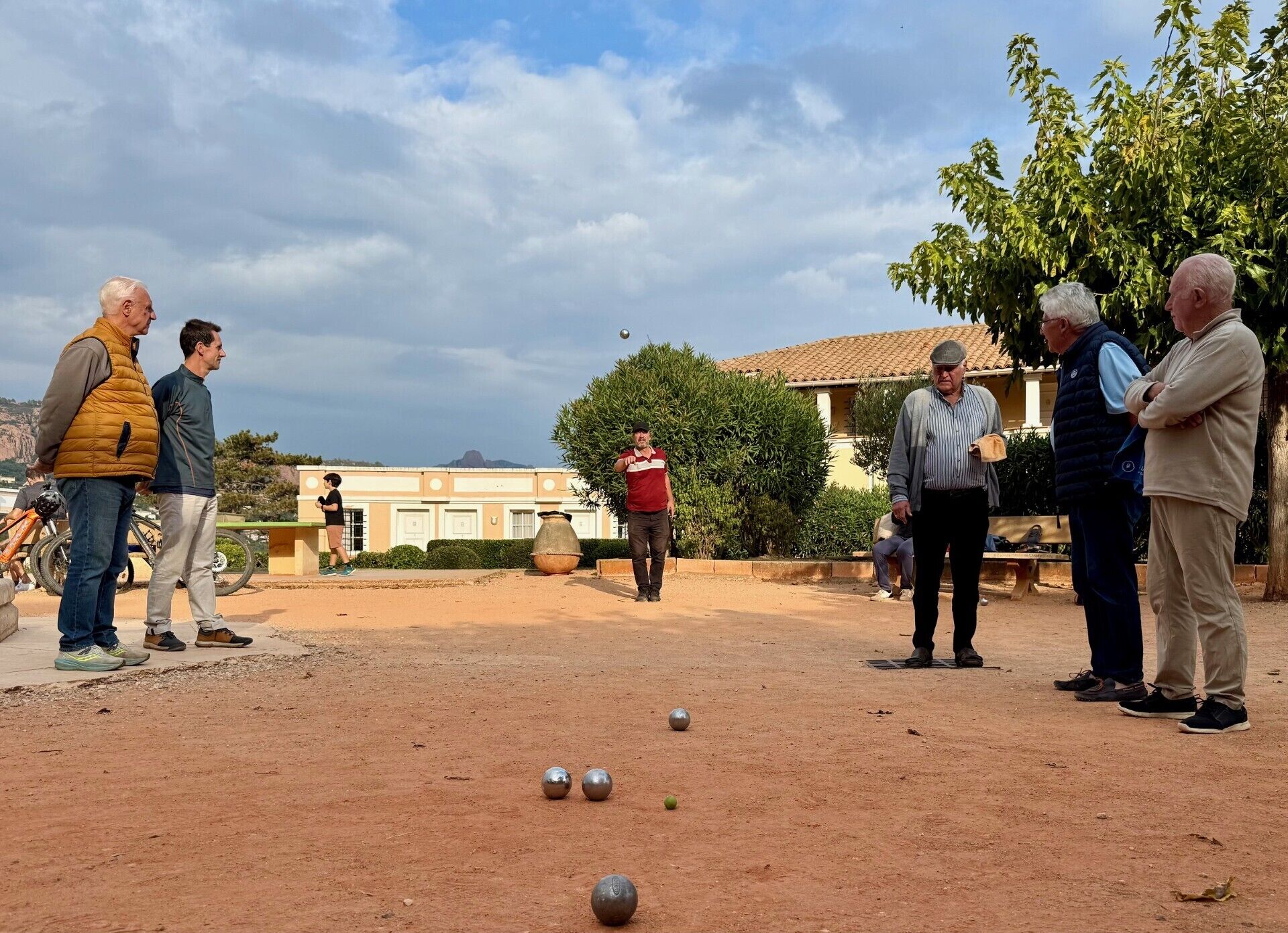 En gruppe mennesker spiller bocce på en jordbane, med en bygning i baggrunden.