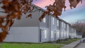 Una hilera de edificios de apartamentos de una planta, de color claro, con ventanas y puertas uniformes, un estacionamiento y un área verde.
