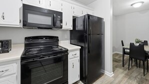 Una cocina con armarios blancos, estufa negra, refrigerador negro y un microondas. En el fondo, se observa una mesa de comedor con sillas.