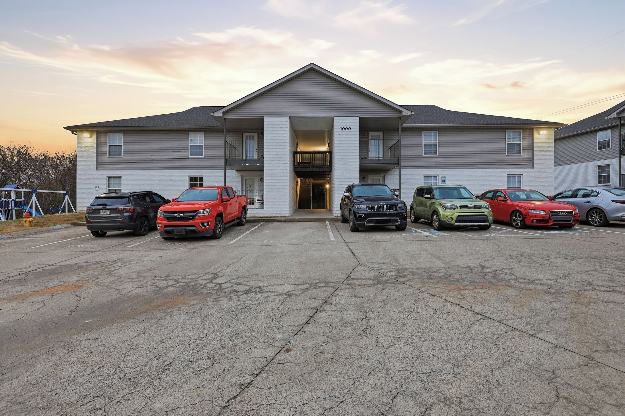 A multi-story building with a central entrance, parking lot with several cars, and a clear sky.