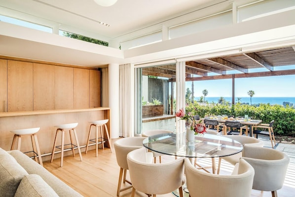 A modern interior with a glass dining table surrounded by chairs, a wooden cabinet, and a view of the ocean. - Elegant Fresnaye Ocean View 4bed Nerine Manor (Cape Town)