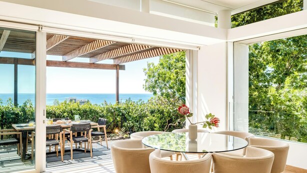 A modern outdoor dining area with a glass table and chairs, overlooking a scenic ocean view. - Elegant Fresnaye Ocean View 4bed Nerine Manor (Cape Town)