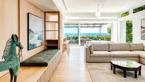 A spacious living room with a large window overlooking the sea, a cozy seating area with a sofa and a coffee table, and a wooden bench with a decorative sculpture. - Elegant Fresnaye Ocean View 4bed Nerine Manor (Cape Town)
