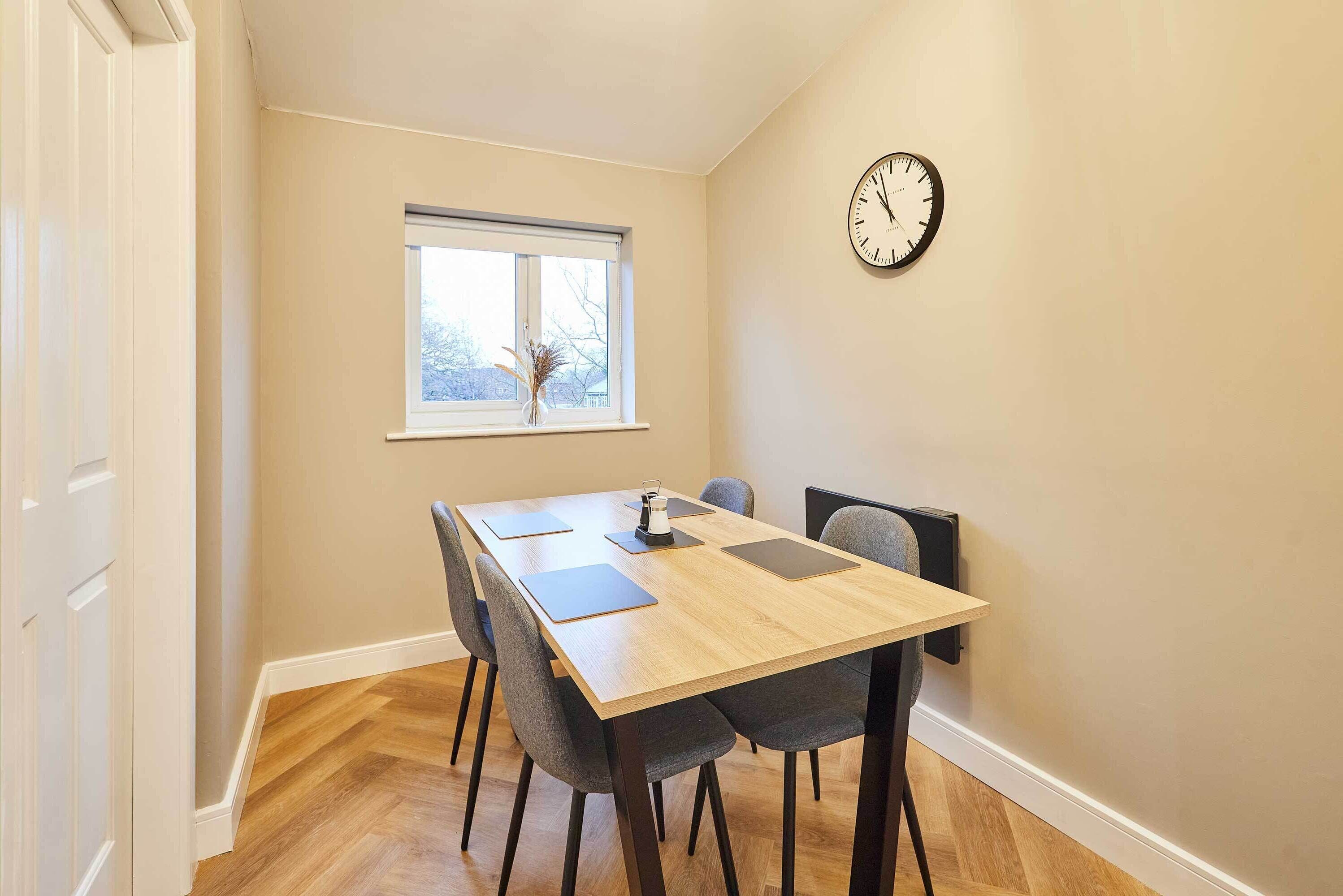 A dining area with a wooden table, four chairs, a clock on the wall, and a window with a view.