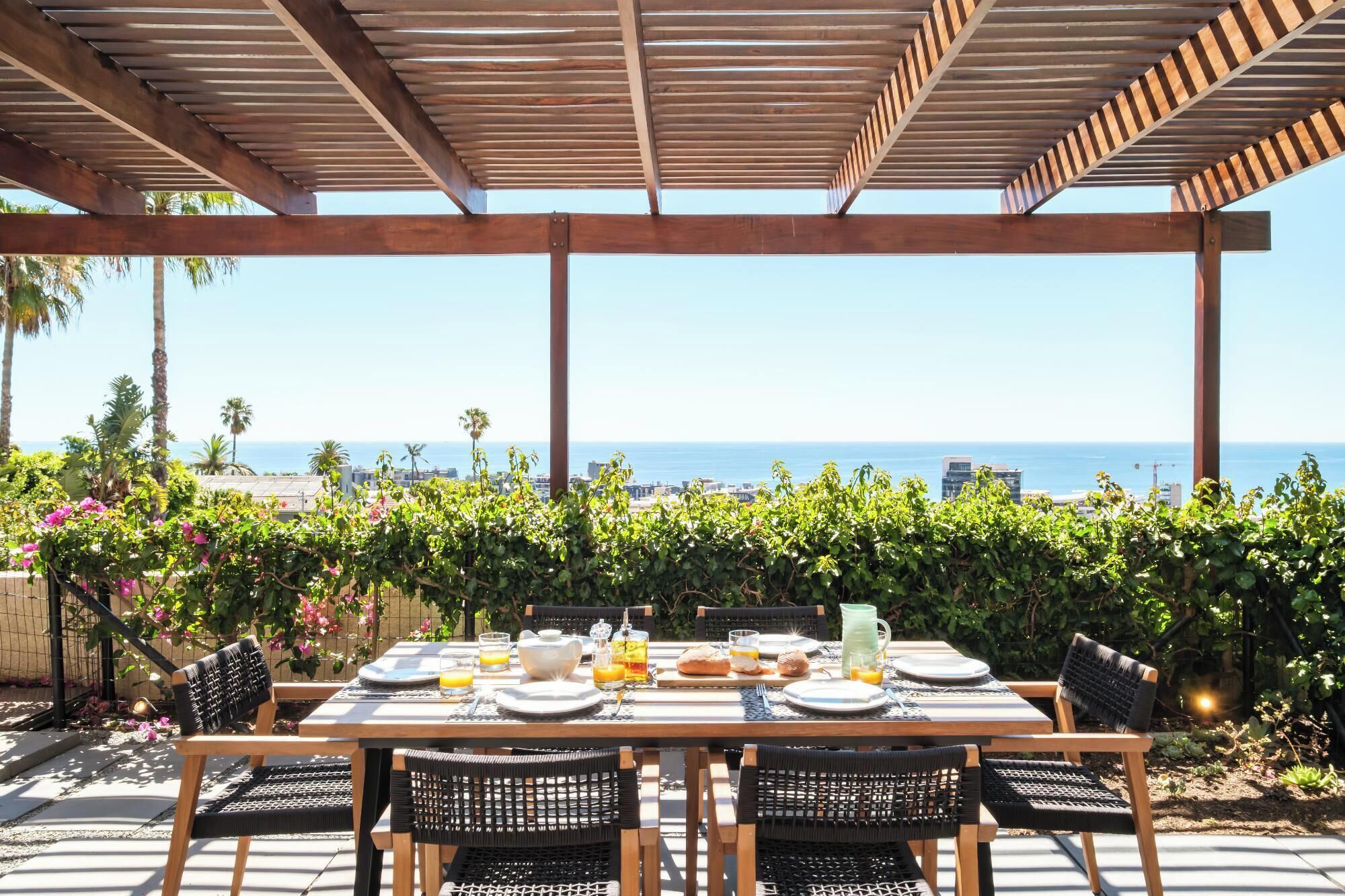 Un área de comedor al aire libre con vistas al océano, pérgola de madera y abundante vegetación.