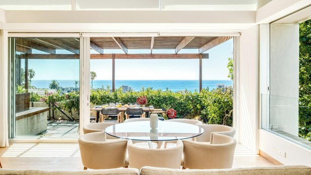 A modern outdoor patio with a glass table and chairs, overlooking a coastal view with a pergola and lush greenery. - Opulent Spacious Fresnaye Sea View Leonotis Villa (Cape Town)