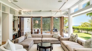 A modern living room with a sectional sofa, a glass coffee table, and a view of a lush garden and ocean. - Opulent Spacious Fresnaye Sea View Leonotis Villa (Cape Town)