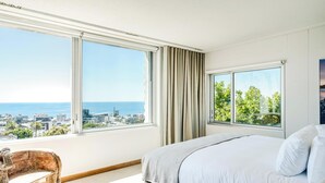 A bedroom with a large bed, a chair, and two windows offering city and ocean views. - Opulent Spacious Fresnaye Sea View Leonotis Villa (Cape Town)