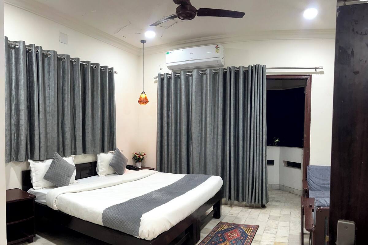 A bedroom with a bed, curtains, a ceiling fan, an air conditioning unit, a nightstand, a phone, a small table with a vase of flowers, and a patterned rug.