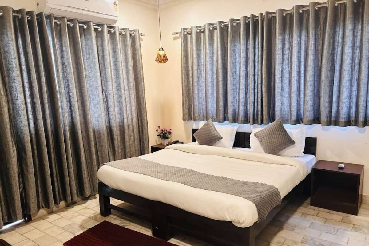A hotel room with a large bed, dark wooden headboard, grey curtains, a red rug, and a small bedside table with a lamp.