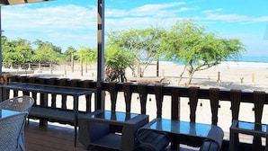 A covered outdoor seating area with wicker furniture and glass tables, overlooking a beach with trees and a clear sky.
