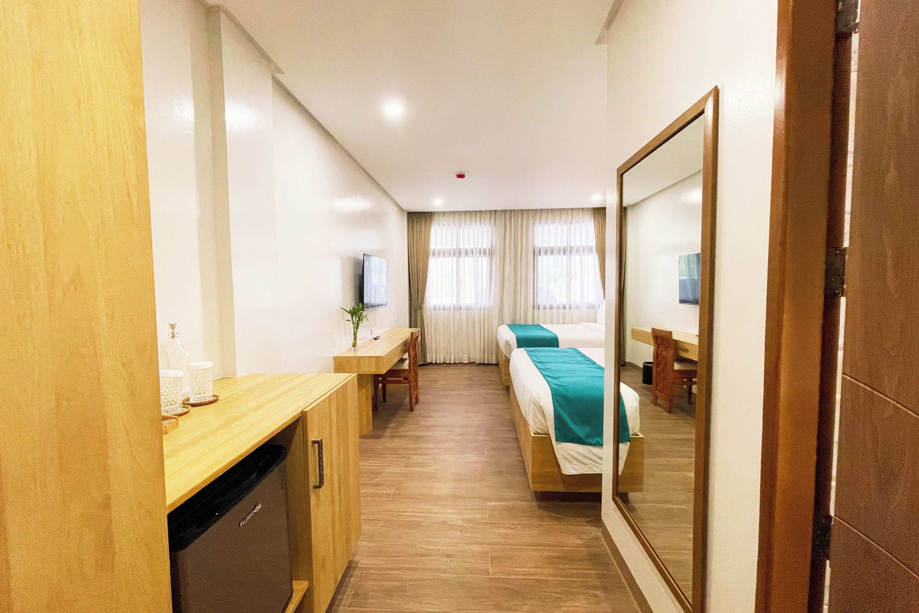 A hotel room with a wooden desk, a bed with a teal bedspread, a mirror, and a television.