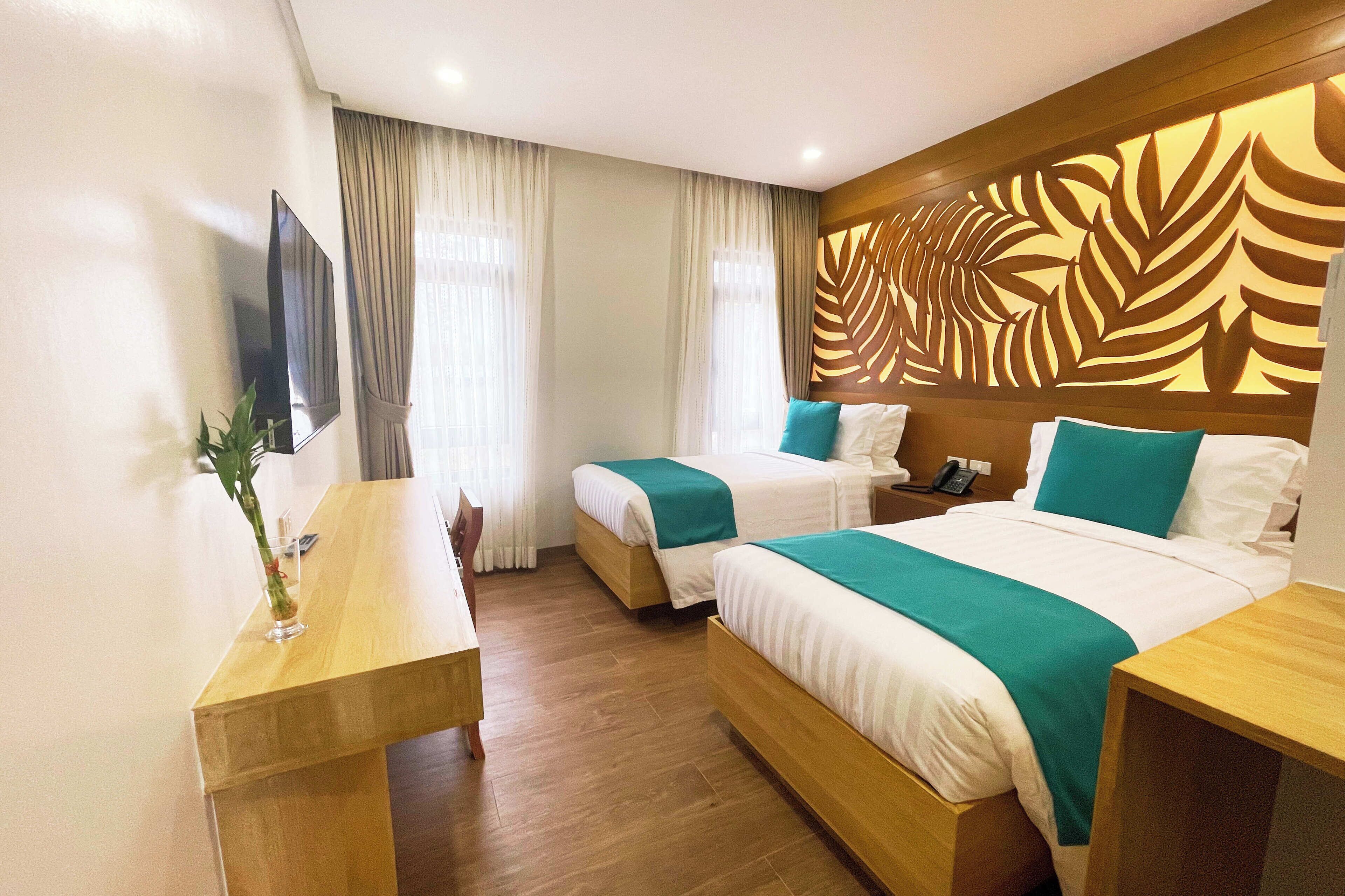 A hotel room with two beds, a wooden table, a TV, and a decorative leaf pattern on the wall.