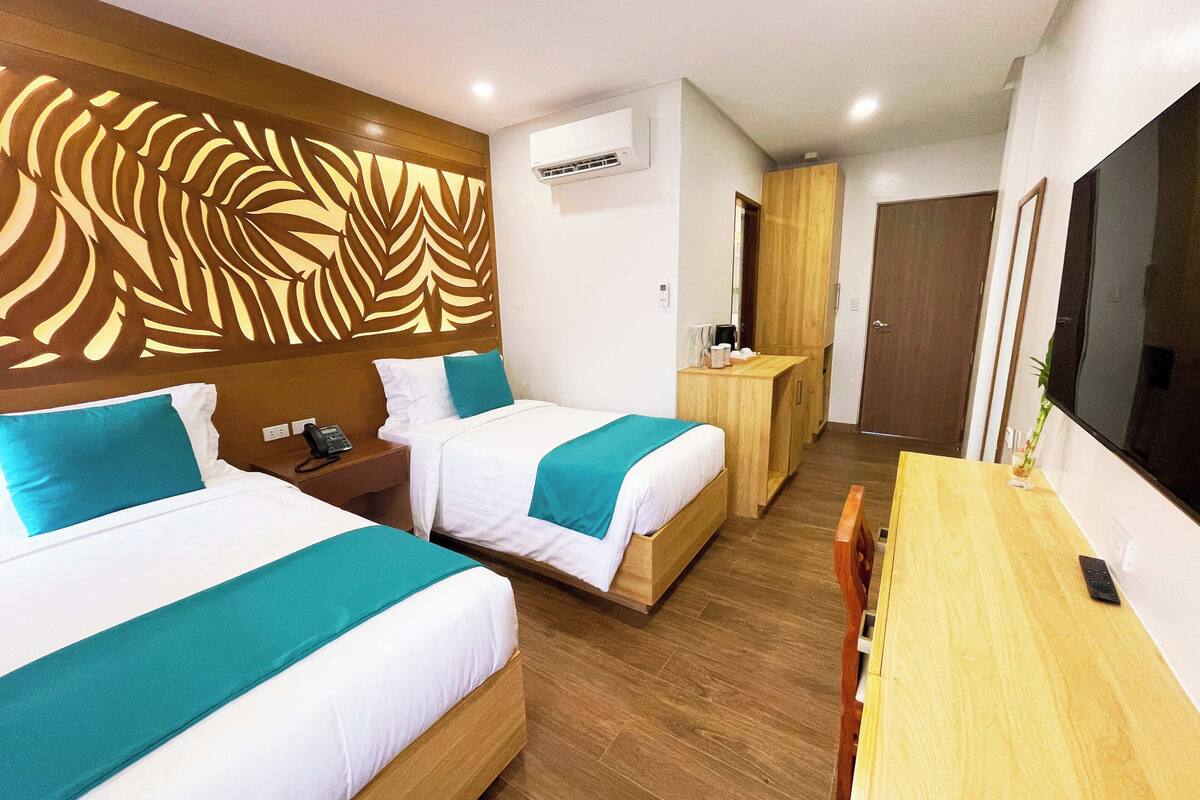 A hotel room with two beds, a wooden desk, a TV, and a wall-mounted air conditioner.
