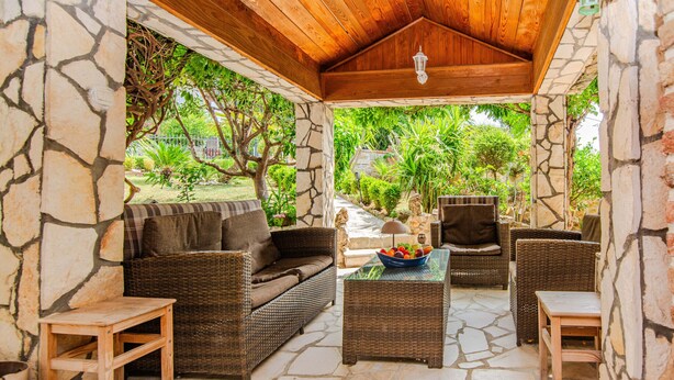 A covered outdoor seating area with wicker furniture, stone walls, and a view of a garden. - Holiday Home Florendale with pool (Maslenica)