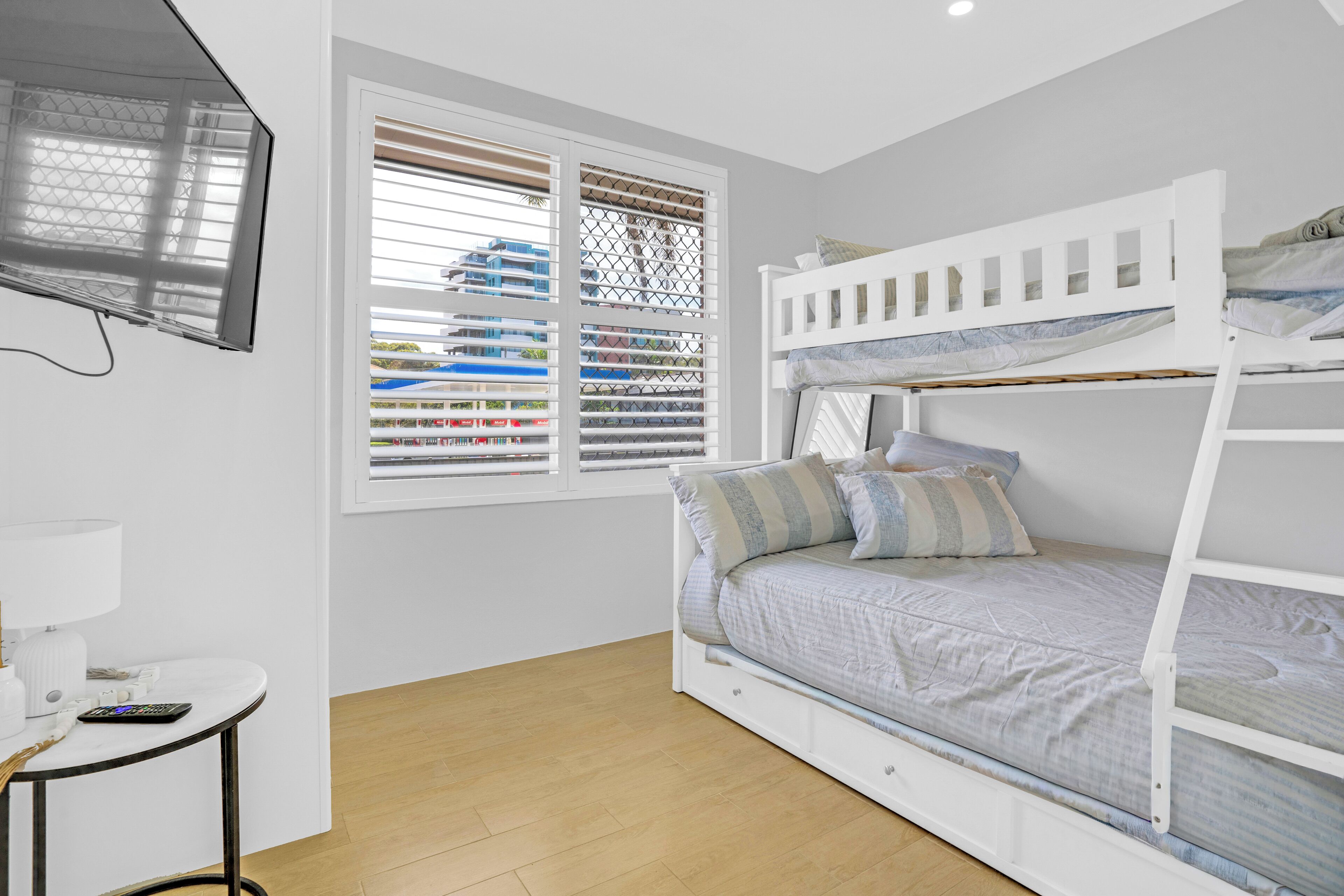 A bunk bed with a TV mounted on the wall, a window with blinds, and a small table with a lamp.
