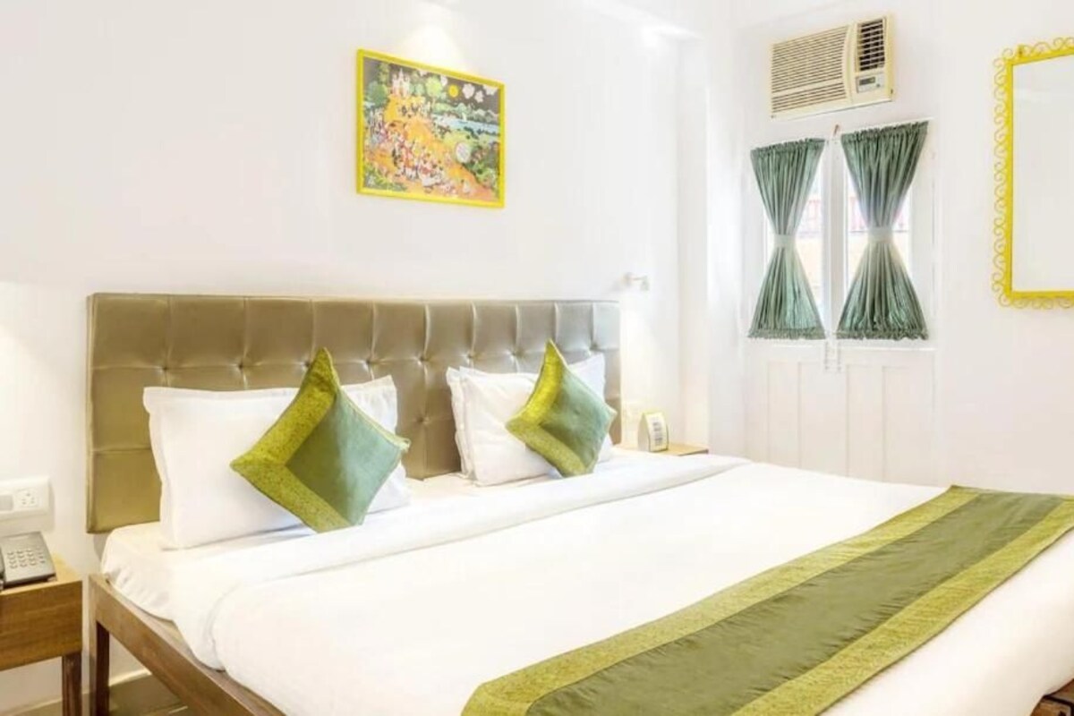 A hotel room with a bed, a bedside telephone, a window with curtains, and a wall-mounted air conditioning unit.