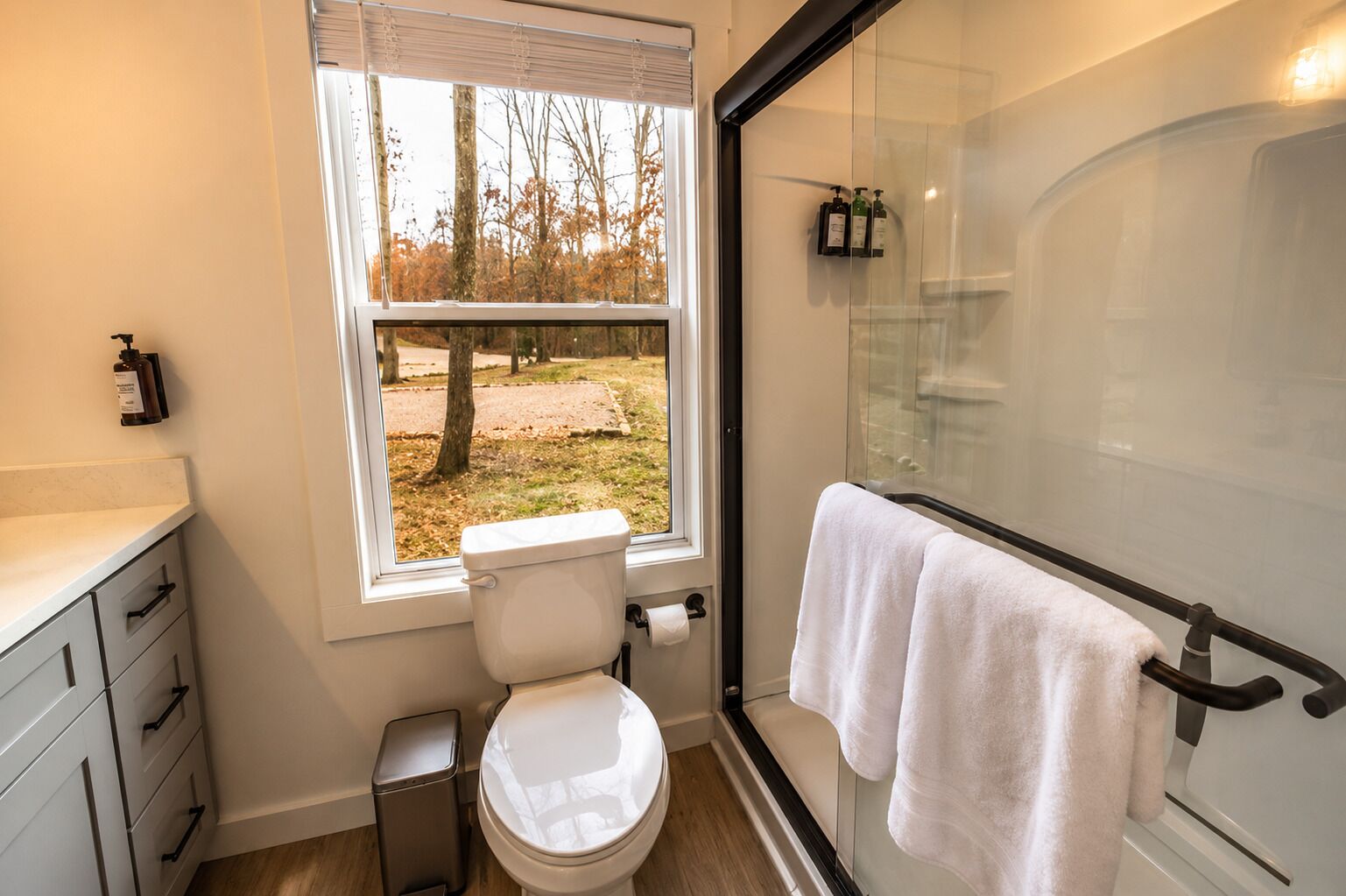 A bathroom with a toilet, a glass shower enclosure, a window with a view of trees, and a towel rack.