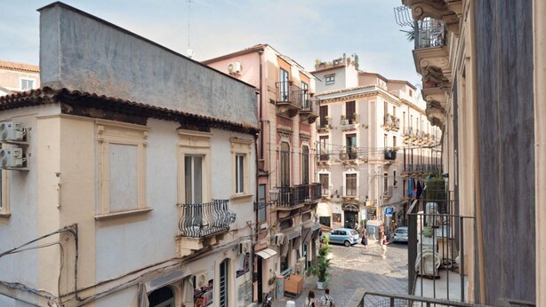 A narrow street with multi-story buildings, balconies, and parked cars. - GuestHost - Charming and cozy 91 square meters apartment, perfect for four people, located on the first floor of the historic Palazzo Auteri Perrotta (NO elevator).Elegantly decorated, it features two frescoed ceilings.The property is located in the hear (Catania)