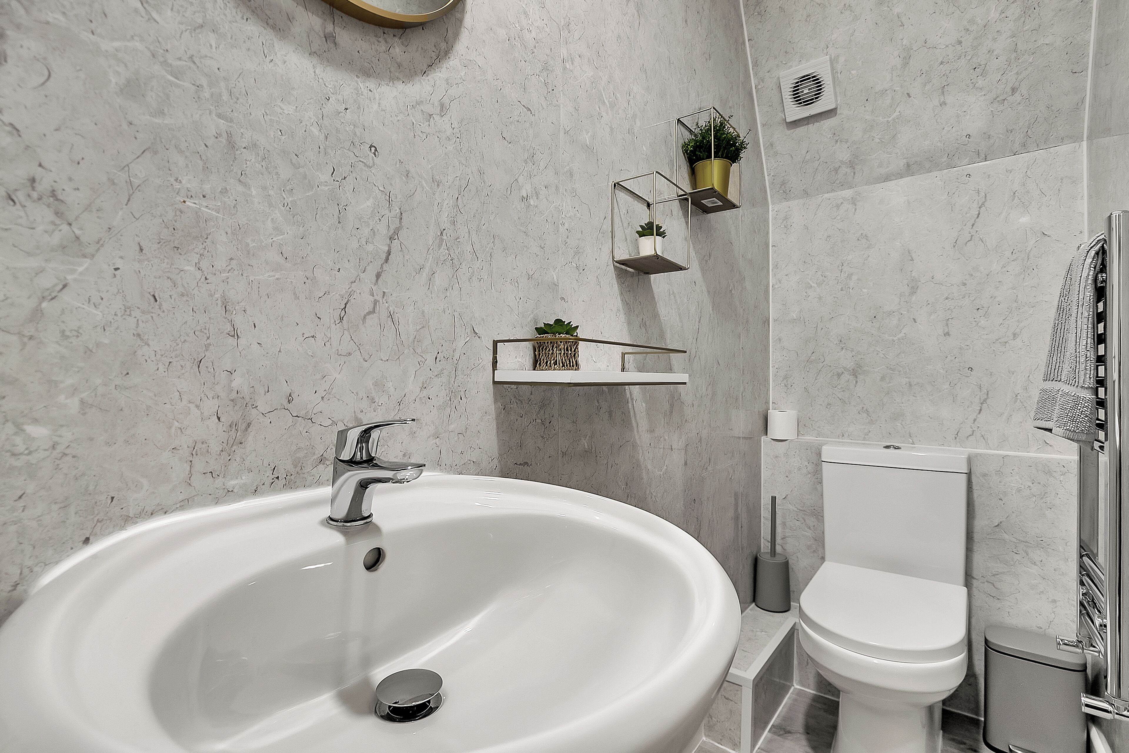 A bathroom with a large white sink, a toilet, and a shower area. There are two wall-mounted shelves with plants.