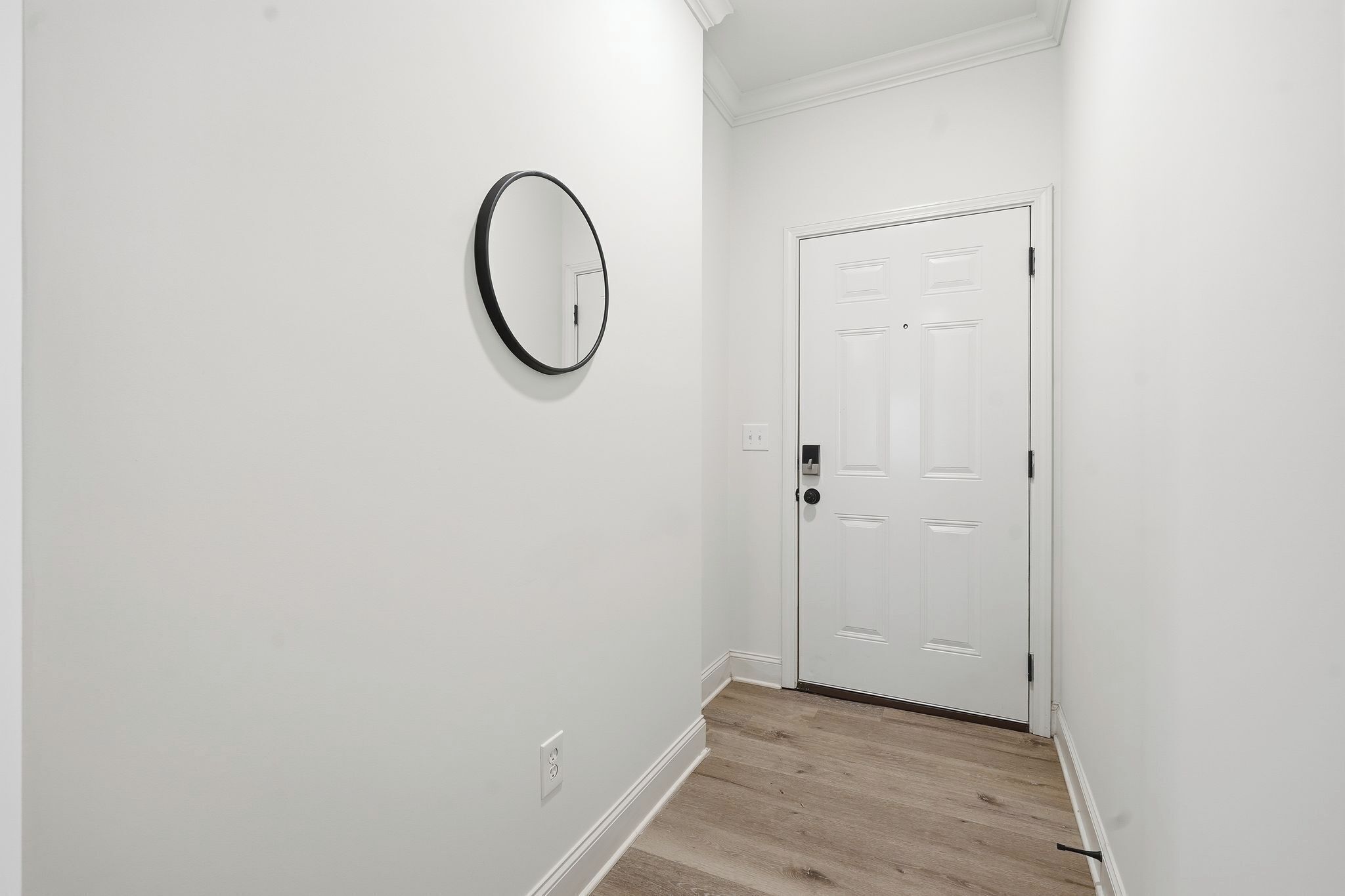 Un pasillo con paredes blancas, piso de madera, una puerta blanca y un espejo redondo en la pared.