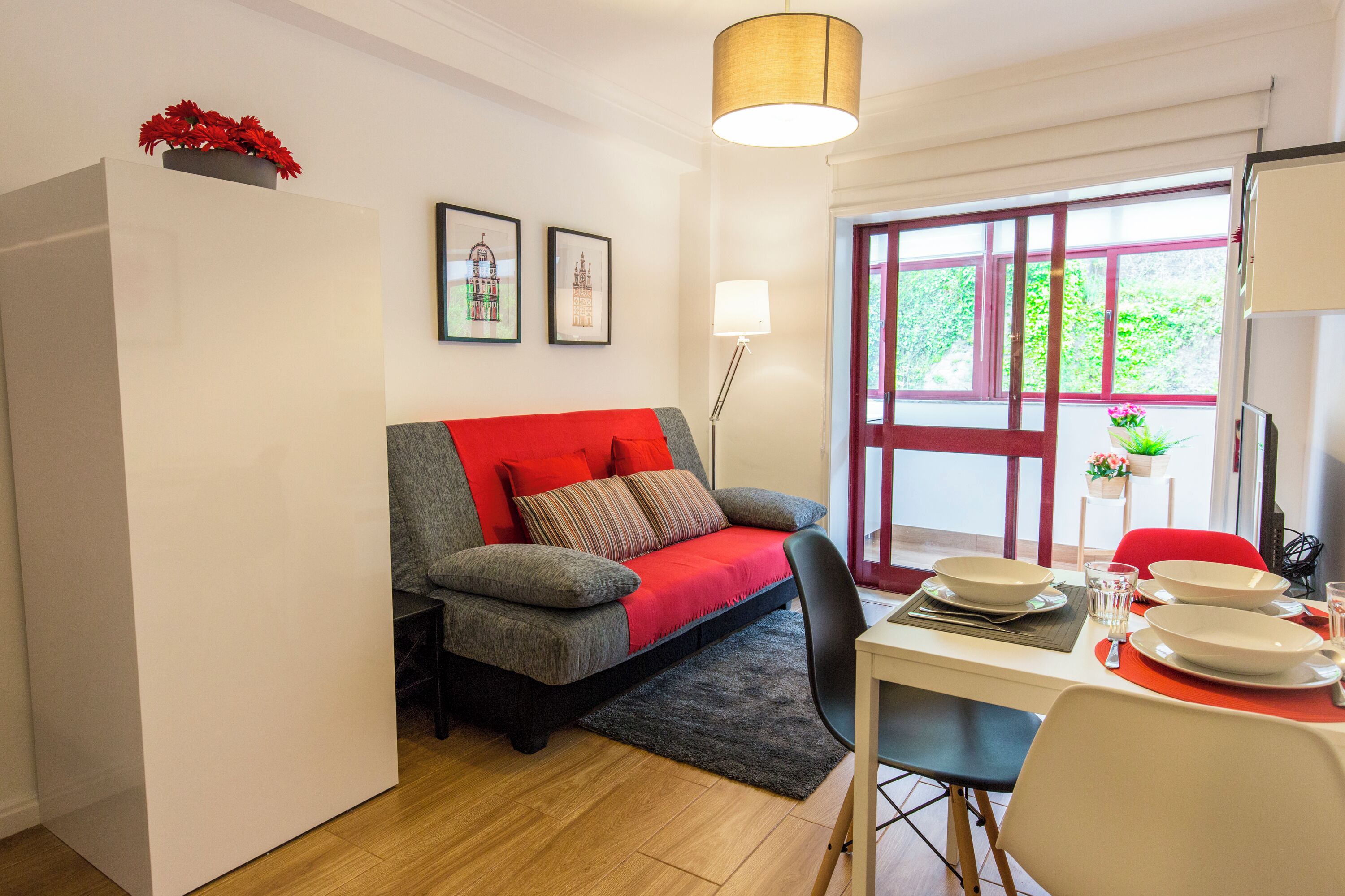 Un salon moderne avec un canapé gris, des coussins rouges, une table à manger avec de l’assiette et des verres, et une grande fenêtre donnant sur un paysage verdoyant.