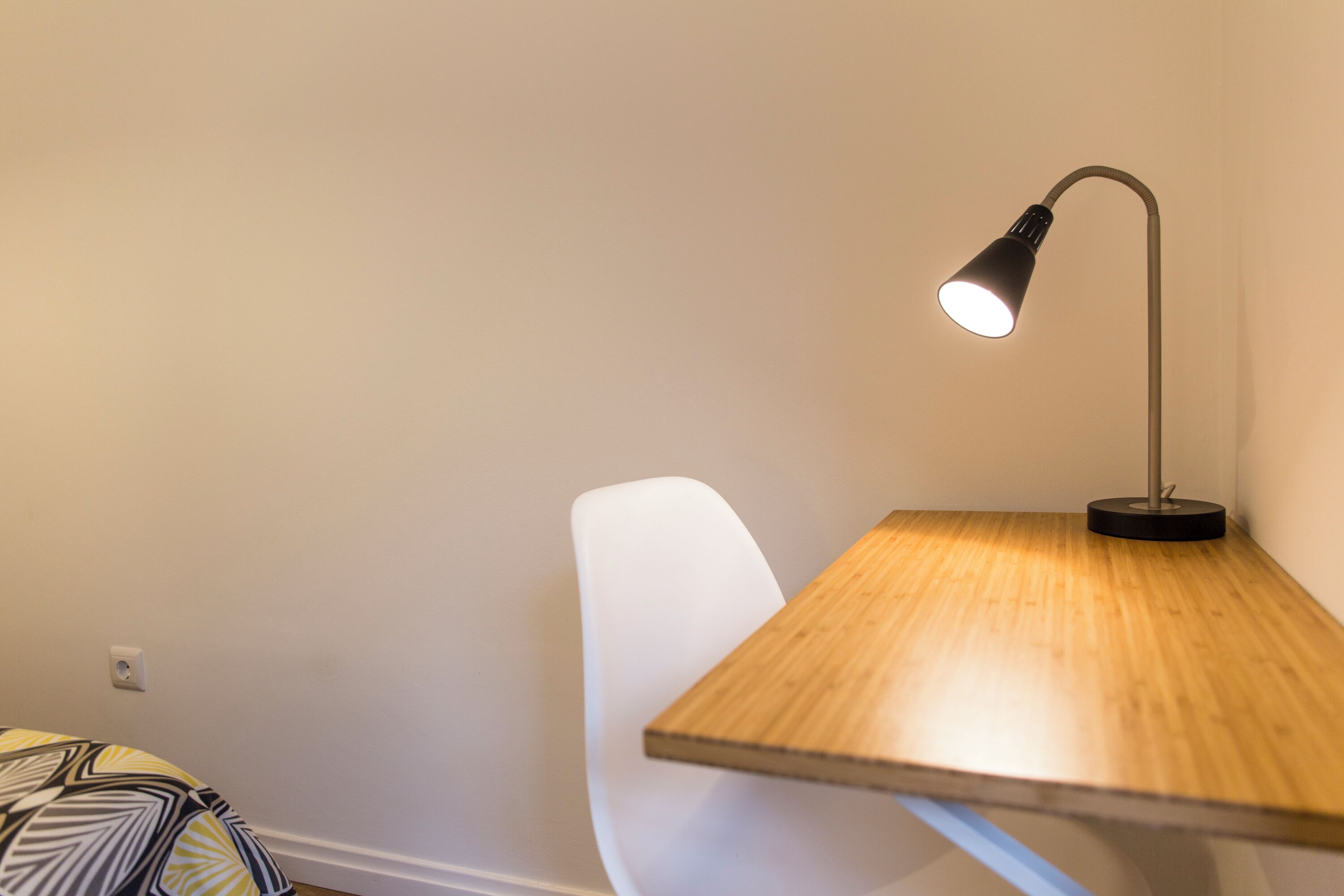 A corner of a room with a wooden table, a white chair, and a desk lamp.