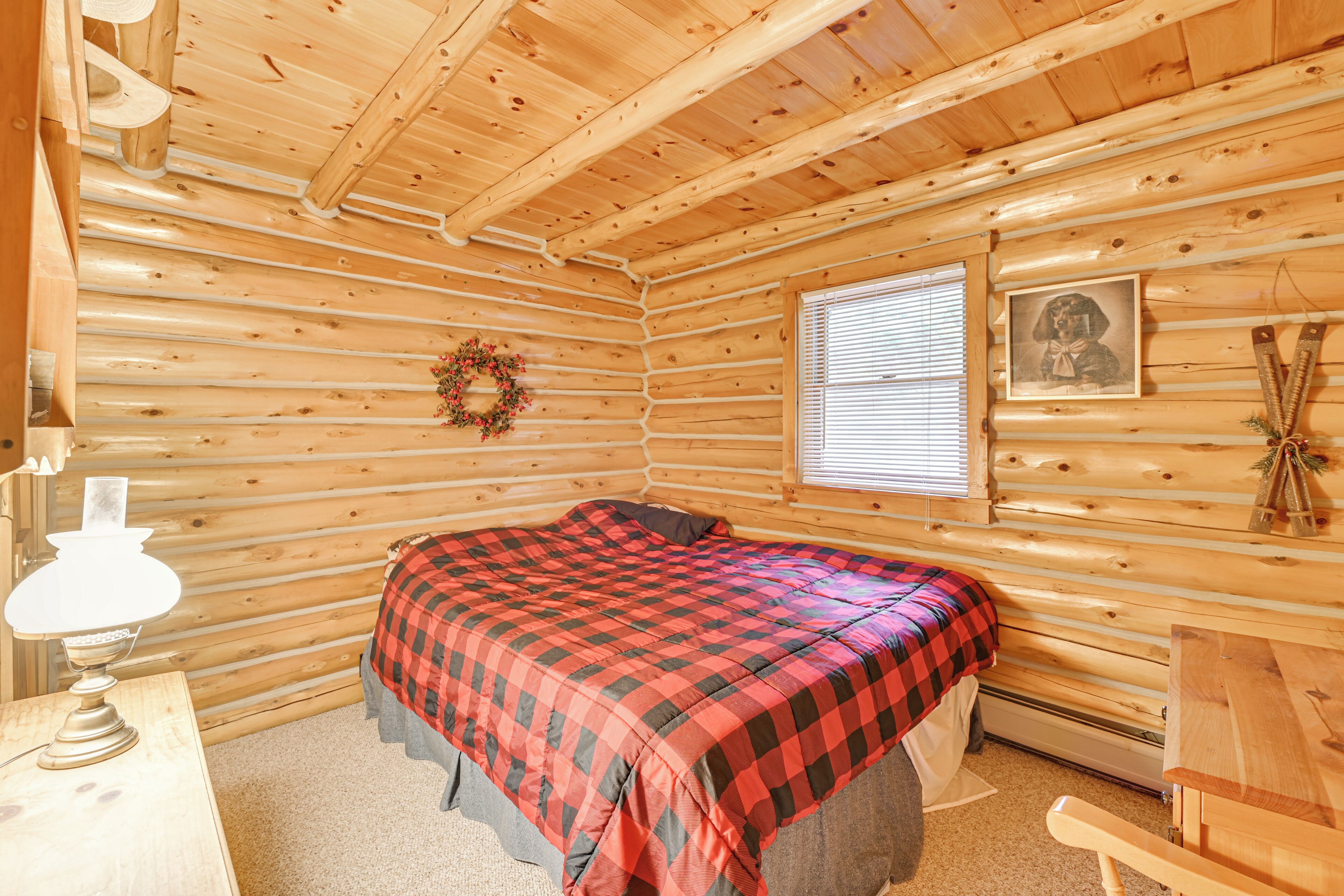 A log cabin bedroom with a bed, a desk with a lamp, a window with blinds, and a framed picture on the wall.