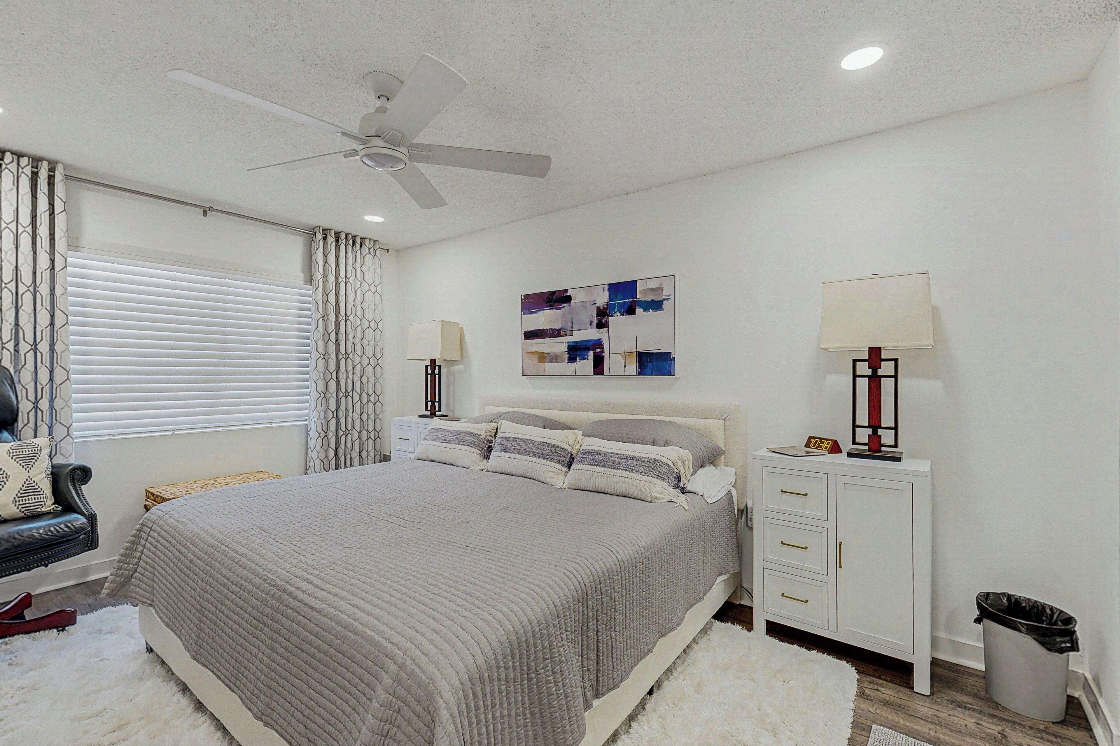 A bedroom with a bed, a ceiling fan, a nightstand with a lamp, a wall art piece, and a trash bin.