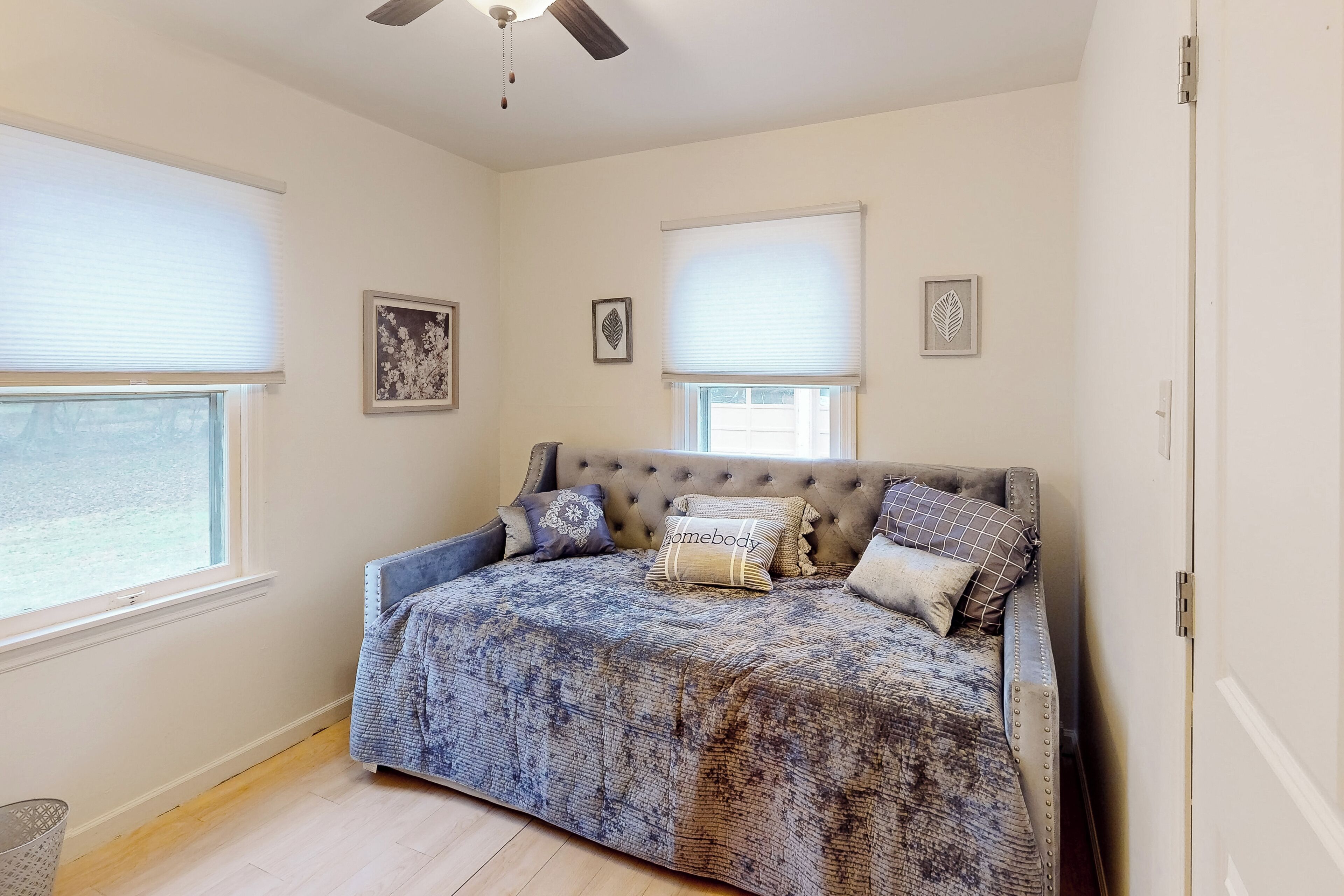 A bedroom with a large bed, two windows with blinds, a ceiling fan, and framed artwork on the walls.