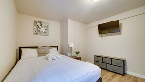 A neatly made bed with a white duvet, a bedside lamp, a small plant, and a TV mounted on the wall. - Walk to Light Rail: Quiet Judkins Park Home Base! (Seattle)