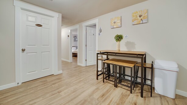 A hallway with a wooden floor, a white door with a sign, a small table with stools, a trash can, and a wall with a flower painting. - Walk to Light Rail: Quiet Judkins Park Home Base! (Seattle)