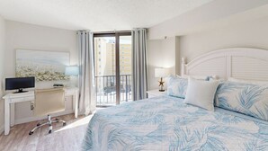 Habitación de hotel con una cama, un escritorio con computadora, una silla, una mesita de noche con lámpara y vista al exterior.
