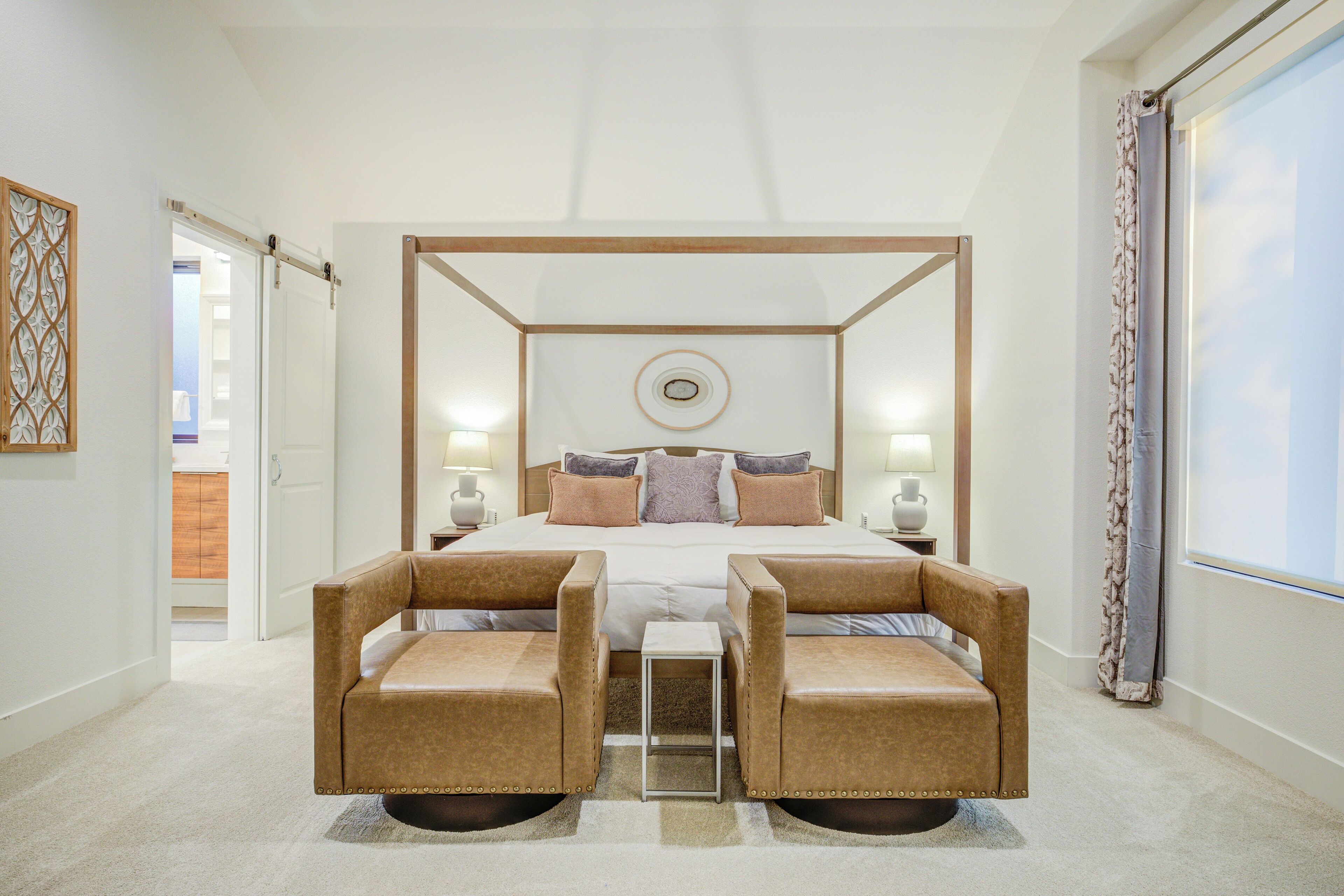 A four-poster bed with a circular wall decoration, two armchairs, a small table, and a view of a room with a wooden door and a window with curtains.