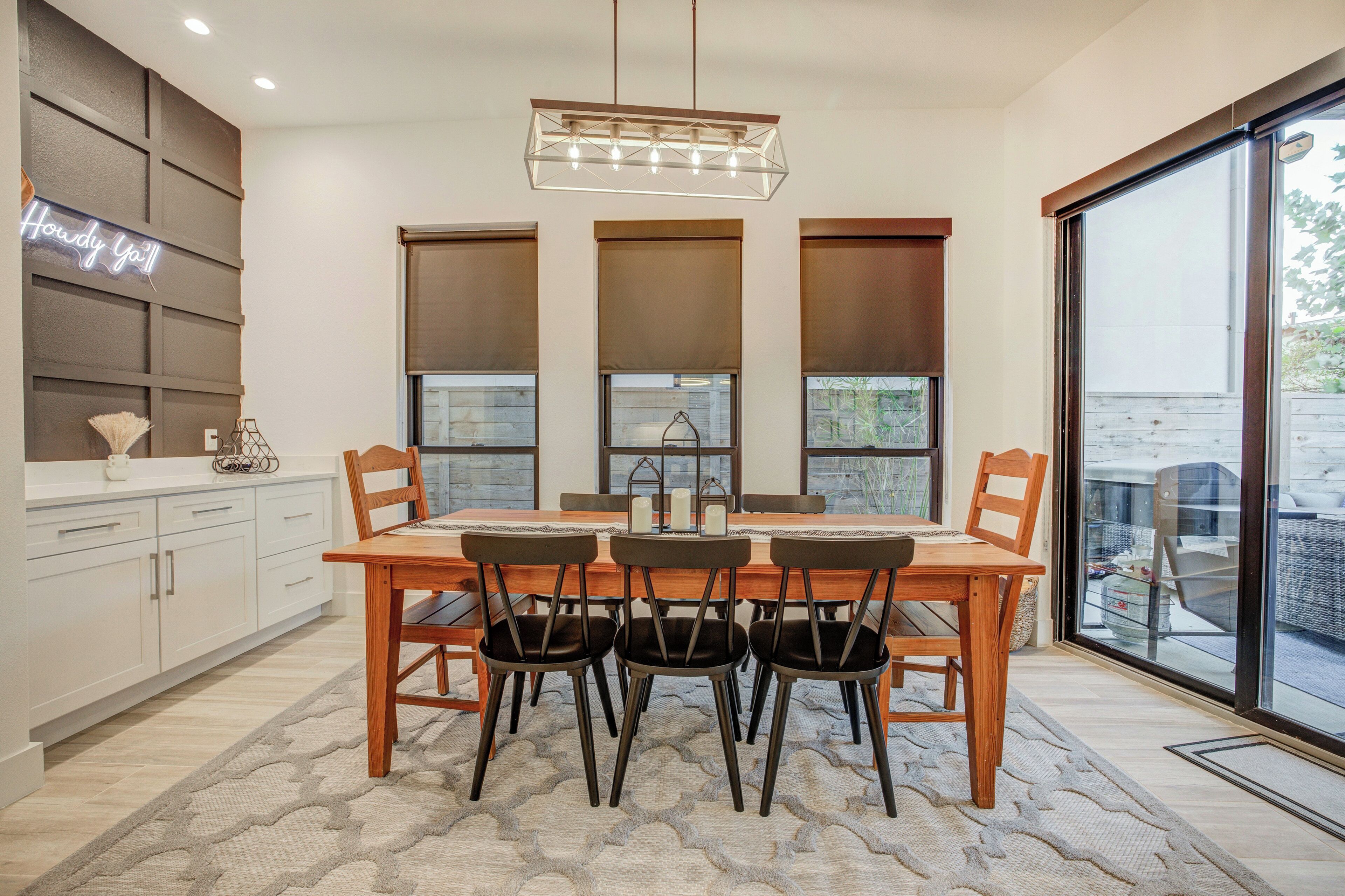 A dining area with a wooden table and chairs, a chandelier, and large windows with blinds.