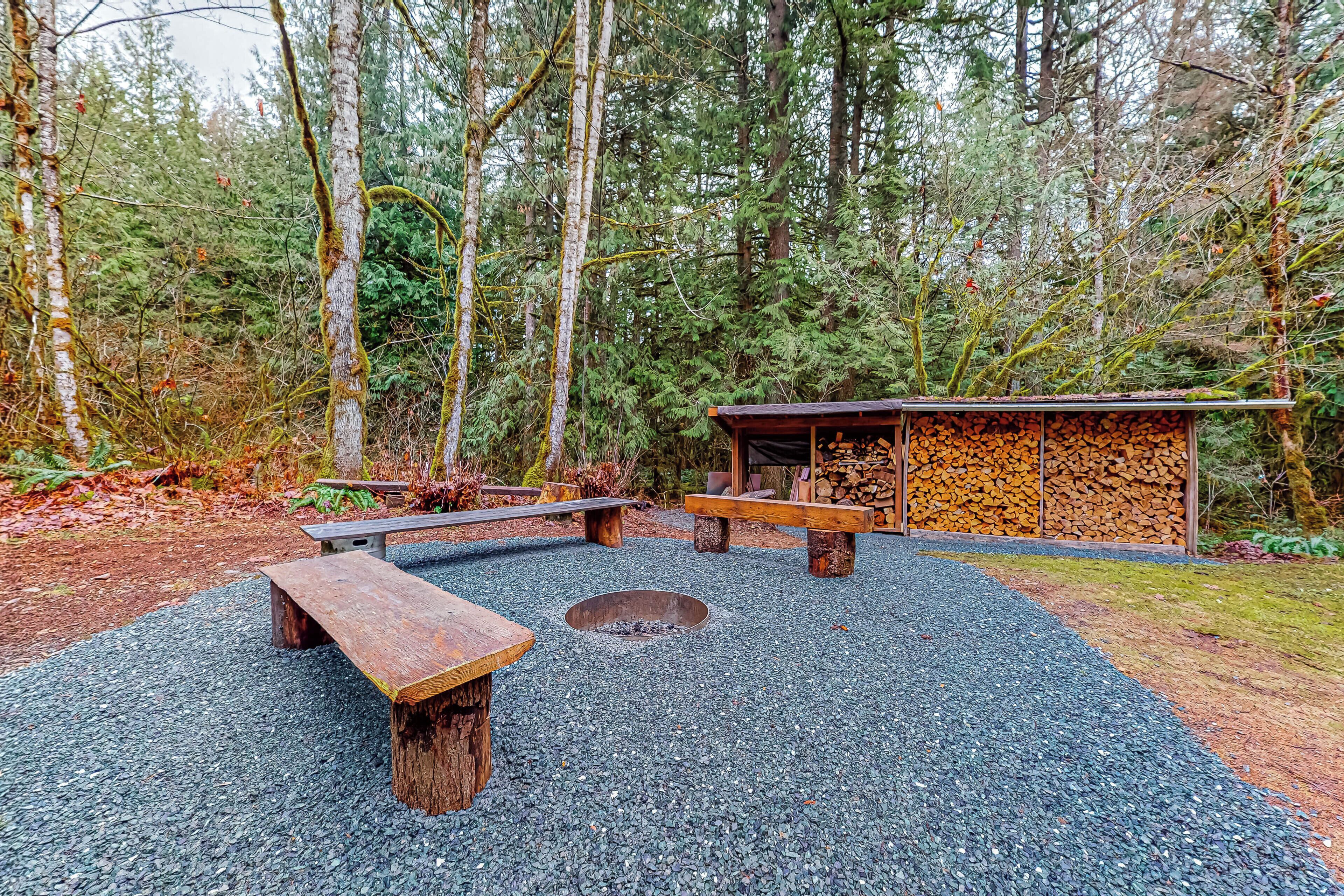 A forested area with a fire pit, benches, and a covered structure with stacked firewood.