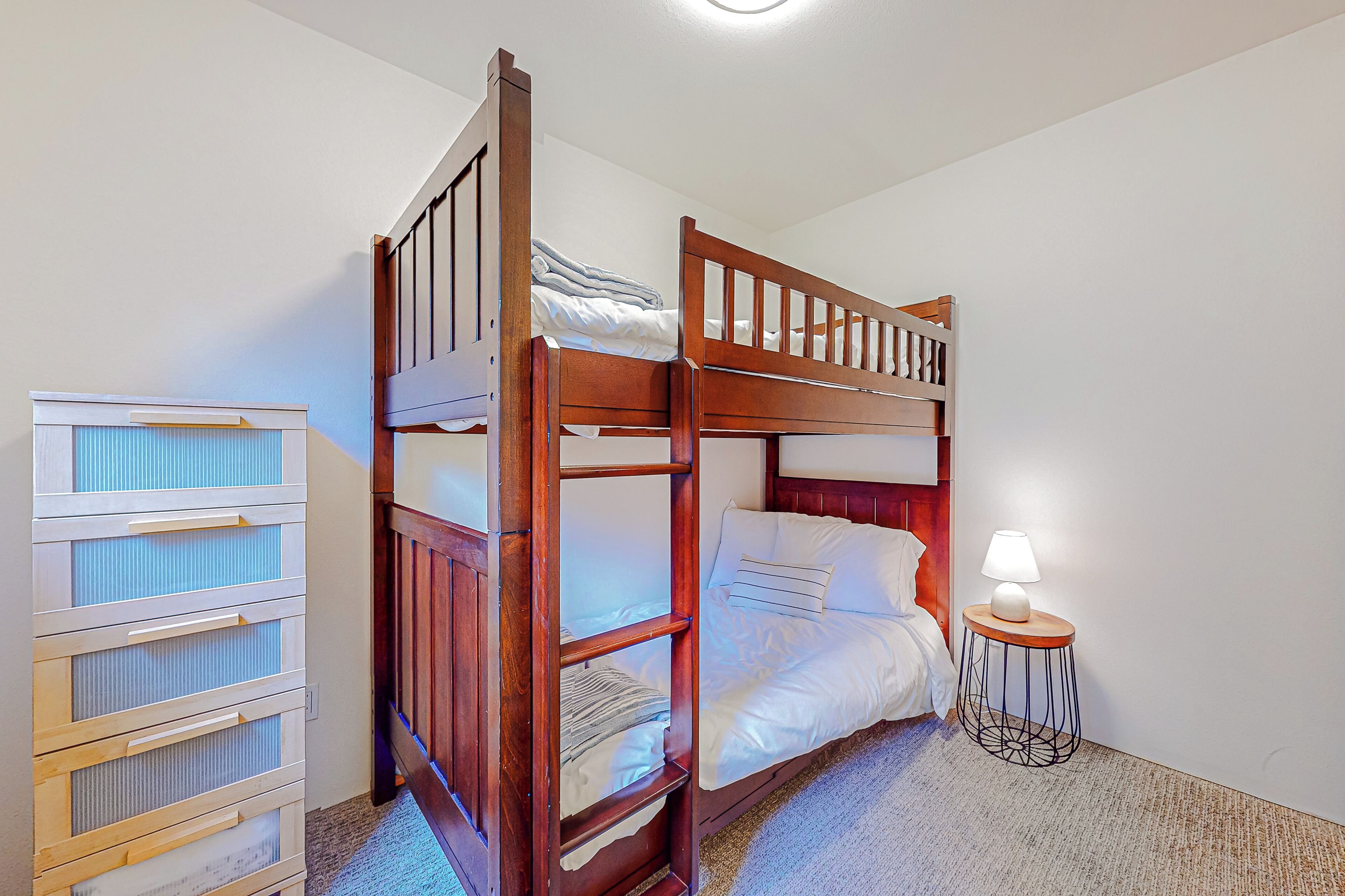 Un lit superposé avec une tête de lit en bois, une table de chevet avec une lampe et une commode avec plusieurs tiroirs.