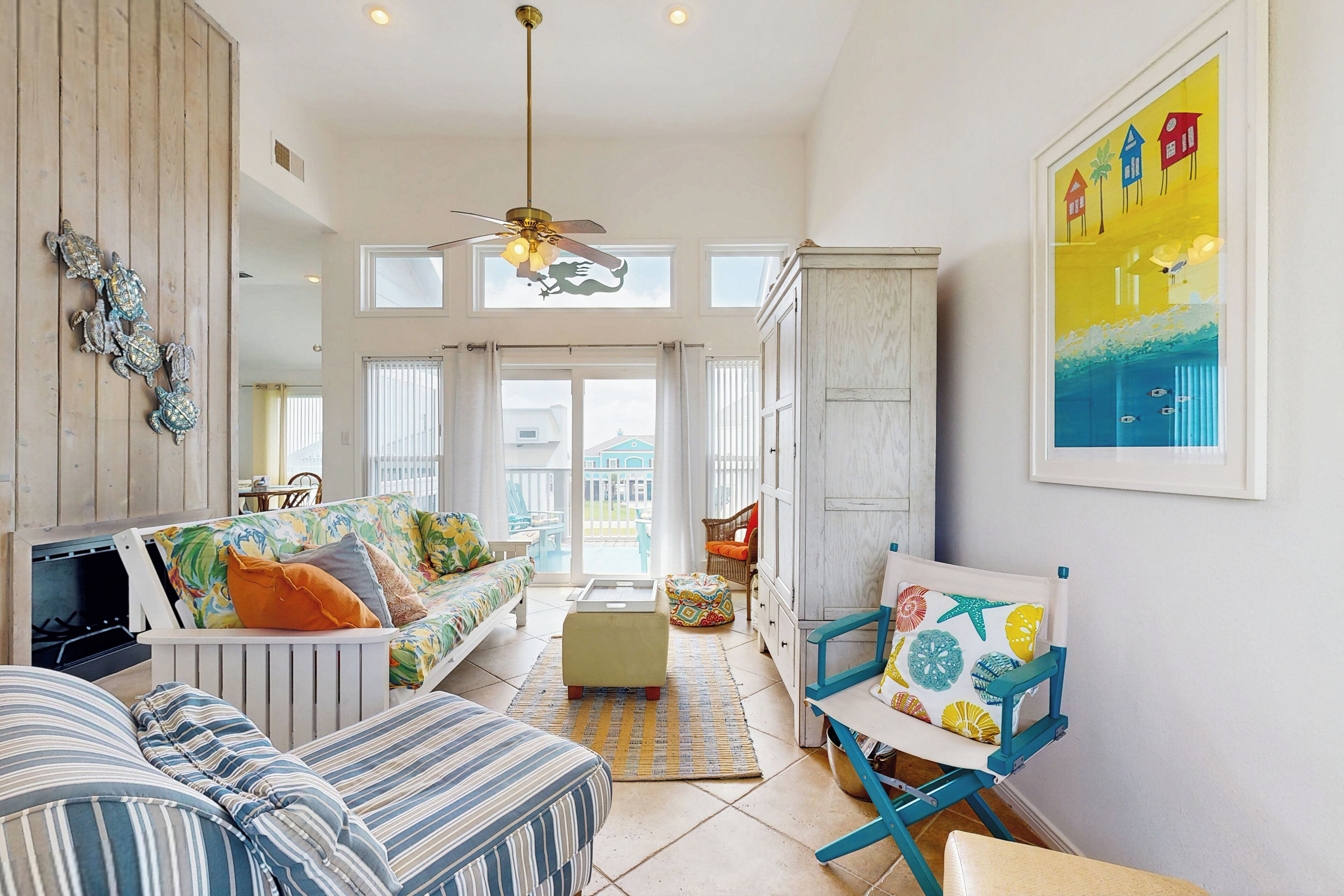 A spacious living area with a high ceiling, a ceiling fan, a sofa with colorful cushions, a wooden cabinet, and a chair with a floral cushion.