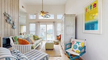 A spacious living area with a high ceiling, a ceiling fan, a sofa with colorful cushions, a wooden cabinet, and a chair with a floral cushion.