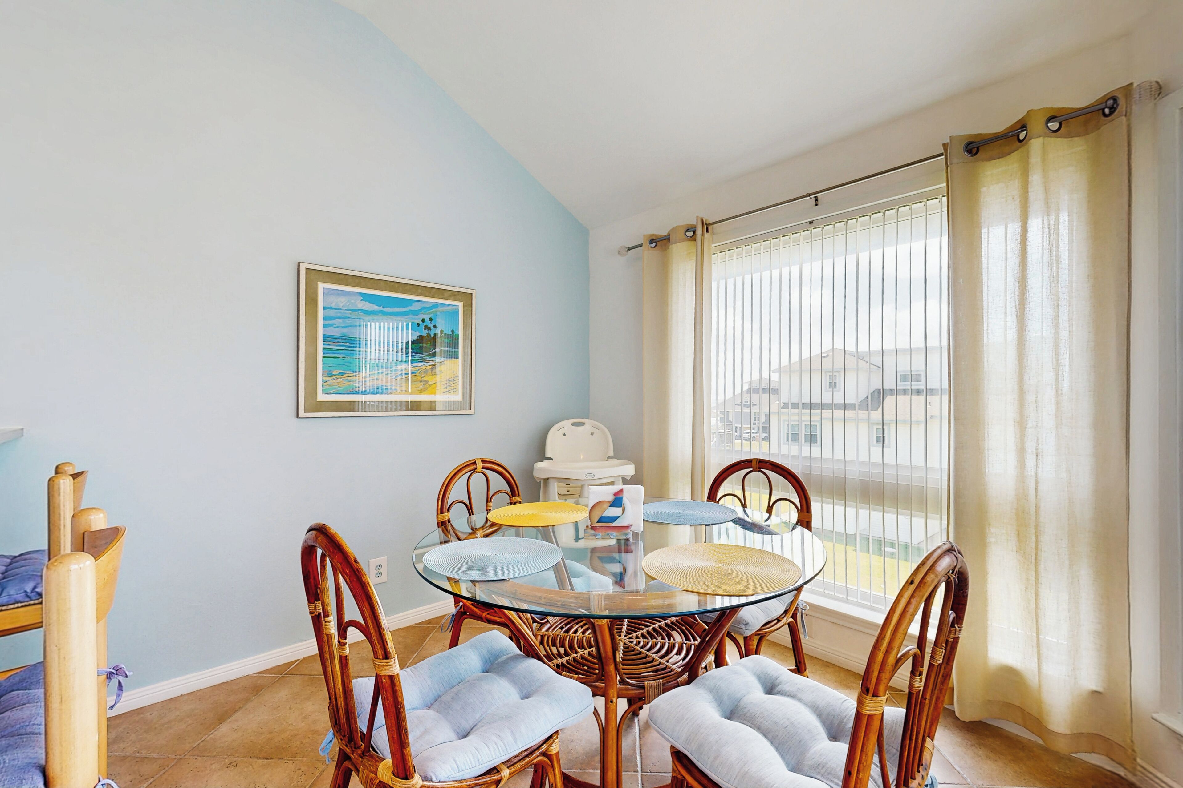 Un comedor con mesa de vidrio rodeada por sillas de mimbre con asientos acolchados, un cuadro en la pared y una ventana con cortinas.