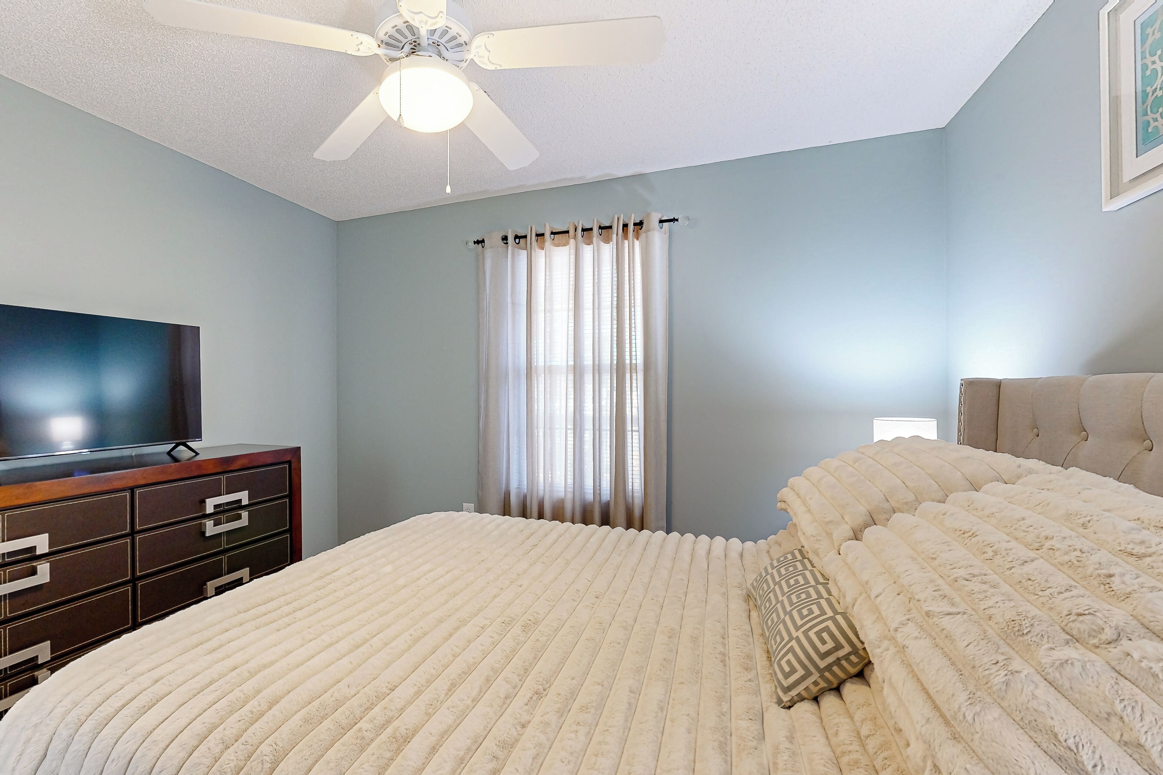 A bedroom with a bed, a TV on a dresser, a ceiling fan, and a window with curtains.